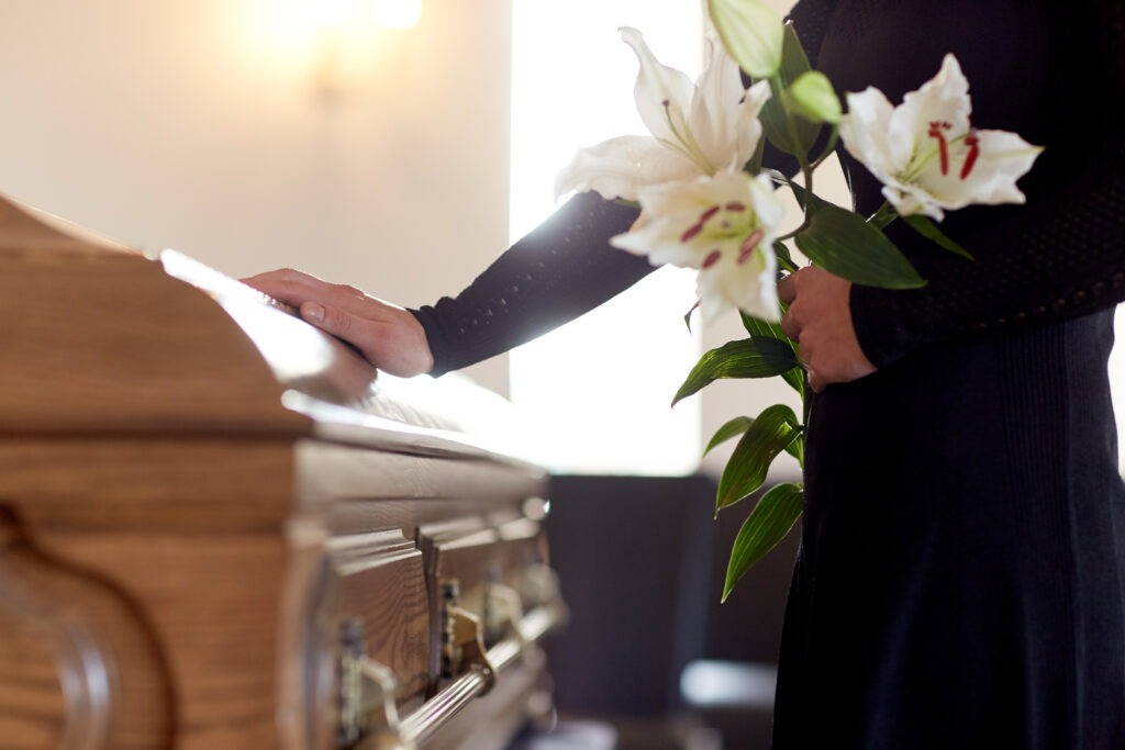 Person placing a hand on a wooden coffin while holding white lilies