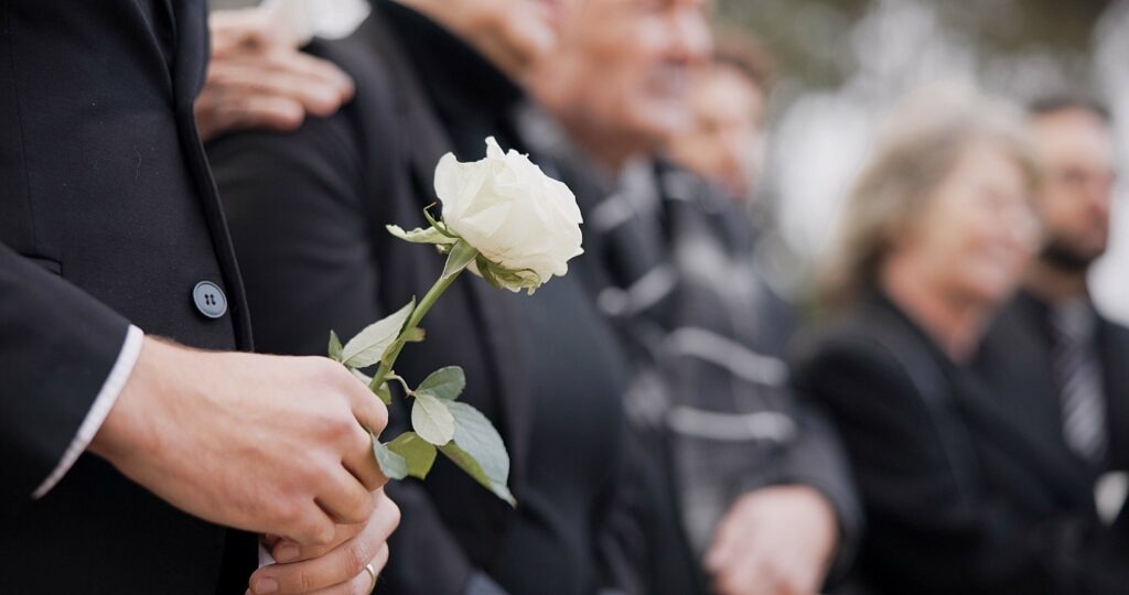 Person holding a white rose during a funeral service with mourners in the background